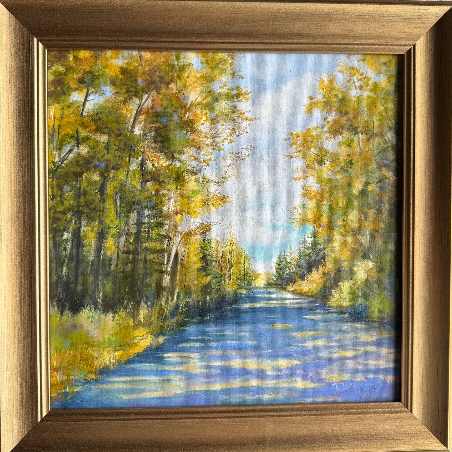 Oil painting of blue sky and a shaded path surrounded by trees with a gold frame. 